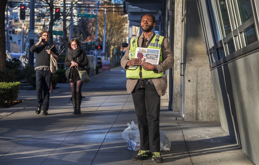 Real Change vendor William Ellington, 25, stands on a sidewalk near an entrance to the Lower Queen Anne QFC, selling papers in 40-degree weather. He keeps possessions in two plastic bags at his feet. He says he hasn’t made much money yet selling the paper, but is making connections he hopes will help in the future. (Credit: Steve Ringman / The Seattle Times)