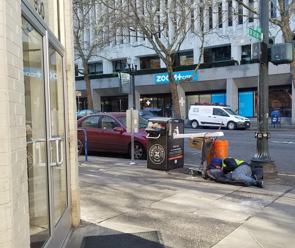 A man sleeps on the sidewalk in front of the 7-Eleven by the Multnomah County Courthouse in January, despite the loud ringing noise radiating from the building. [Credit: Street Roots]