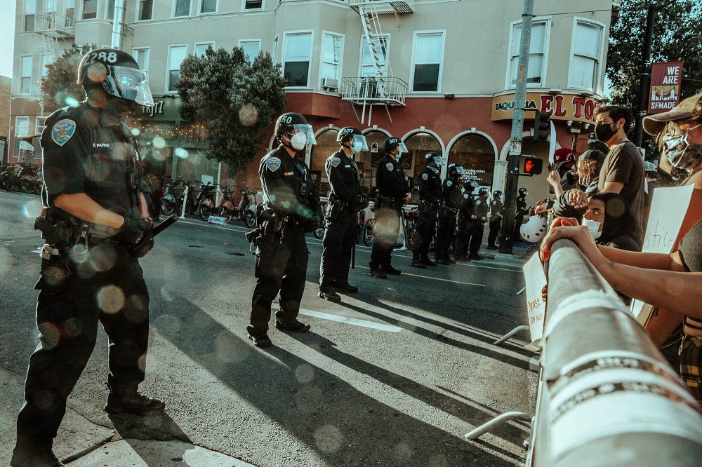 A line of police stand behind a barrier in front of protesters at the Mission police station. [Credit: Kit Castagne]