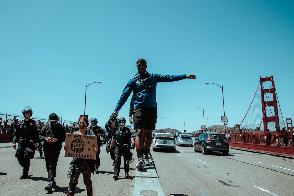 After news breaks that a woman in labor is stuck in the halted Golden Gate Bridge traffic, a protester with a megaphone asks marchers to unblock the road. [Credit: Kit Castagne]